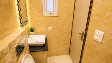A bathroom with a toilet, sink, and shower. There is a window above the sink and a door to the right.
