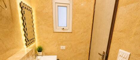 Un baño con inodoro, lavamanos y ducha. Hay una ventana sobre el lavamanos y una puerta a la derecha.