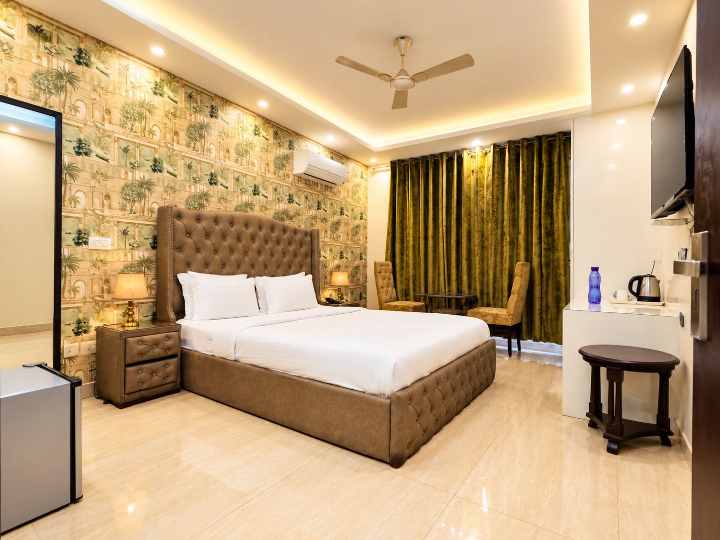 A hotel room with a large bed, a desk, a chair, a small table, a ceiling fan, and a wall-mounted TV.
