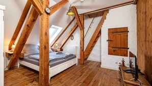 A loft bedroom with a sloped ceiling, wooden beams, a bed, a desk with a lamp, and a window.