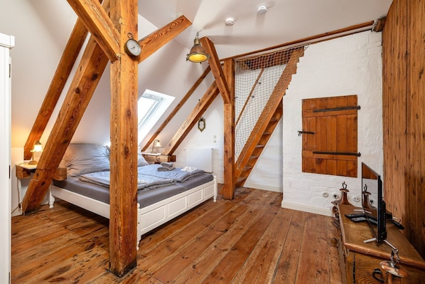 A loft bedroom with a sloped ceiling, wooden beams, a bed, a desk with a lamp, and a window.