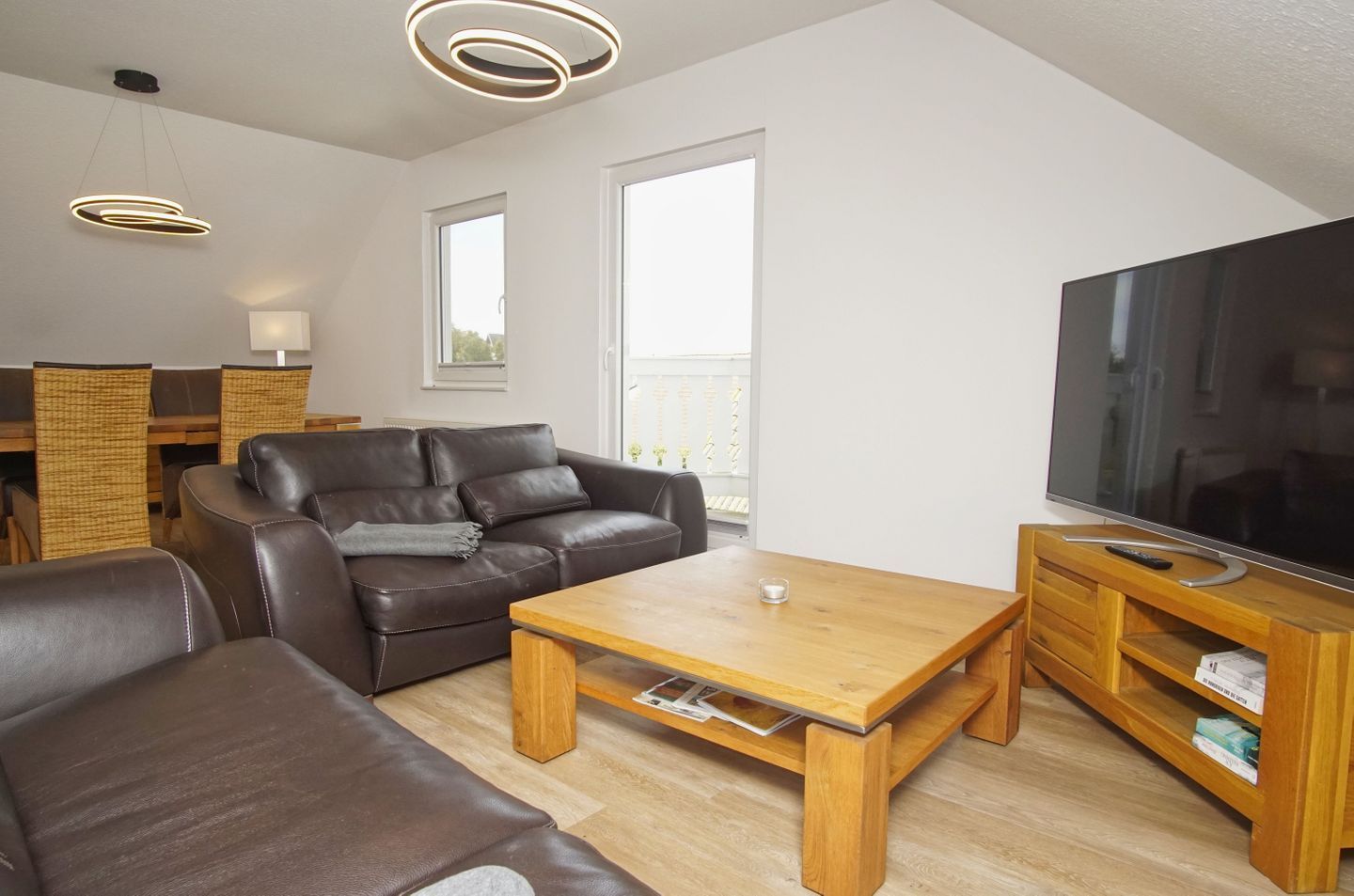 A modern living room with a leather sofa, a wooden coffee table, a TV on a wooden stand, and a balcony visible through the window.