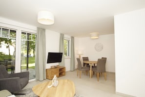 A modern living room with a flat-screen TV, a wooden coffee table, a sofa, and a dining area with chairs and a table. - Beachfront vacation rental with a terrace, pool access, and a sauna - Rex Rugia - Rex (Göhren)