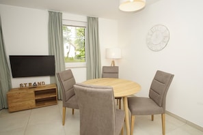 A modern living room with a round wooden table, four grey chairs, a flat-screen TV, a wooden cabinet, a lamp, and a wall clock. - Beachfront vacation rental with a terrace, pool access, and a sauna - Rex Rugia - Rex (Göhren)
