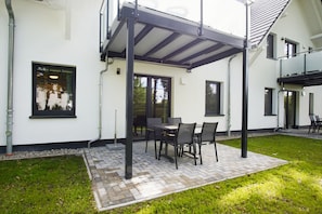 A covered patio area with a table and chairs, a brick pathway, and a white building with windows. - Beachfront vacation rental with a terrace, pool access, and a sauna - Rex Rugia - Rex (Göhren)