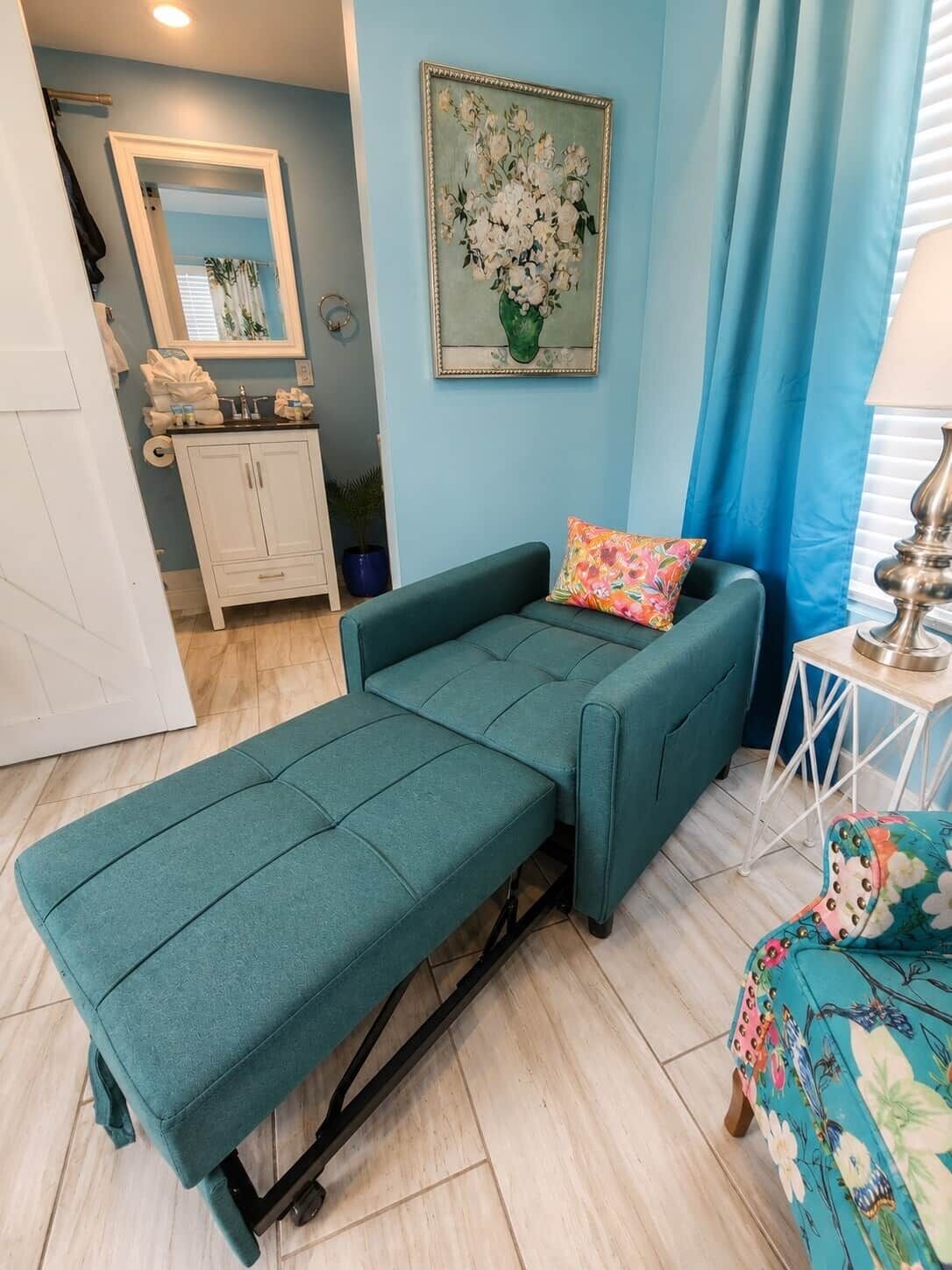 Un canapé de couleur turquoise agrémenté d’un coussin aux couleurs vives, un tableau représentant des fleurs et une petite table avec une lampe.