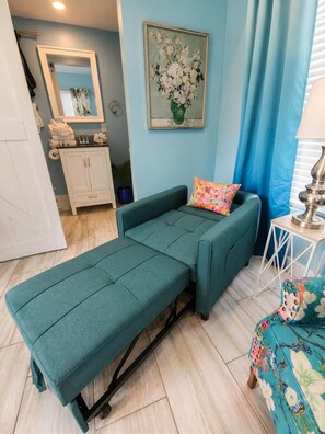 Ein blaugrünes Sofa mit farbenfrohem Kissen, ein Blumenbild und ein kleiner Tisch mit Lampe.
