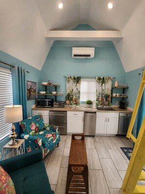 Una cocina compacta con microondas, tostadora y lavabo, junto a un comedor con sofá floral y banco de madera.