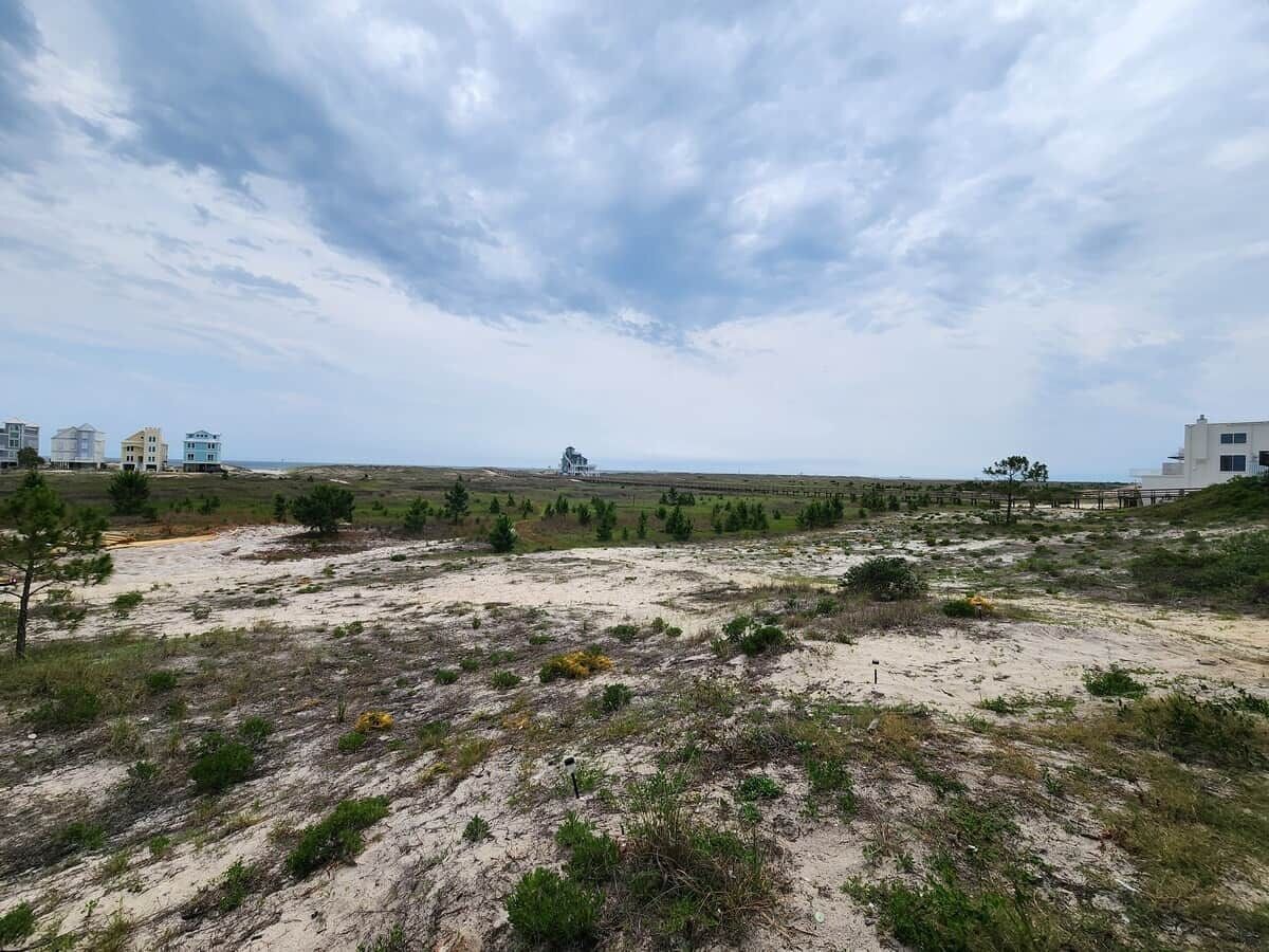 A coastal landscape with sandy terrain and sparse vegetation, featuring a few buildings in the distance.