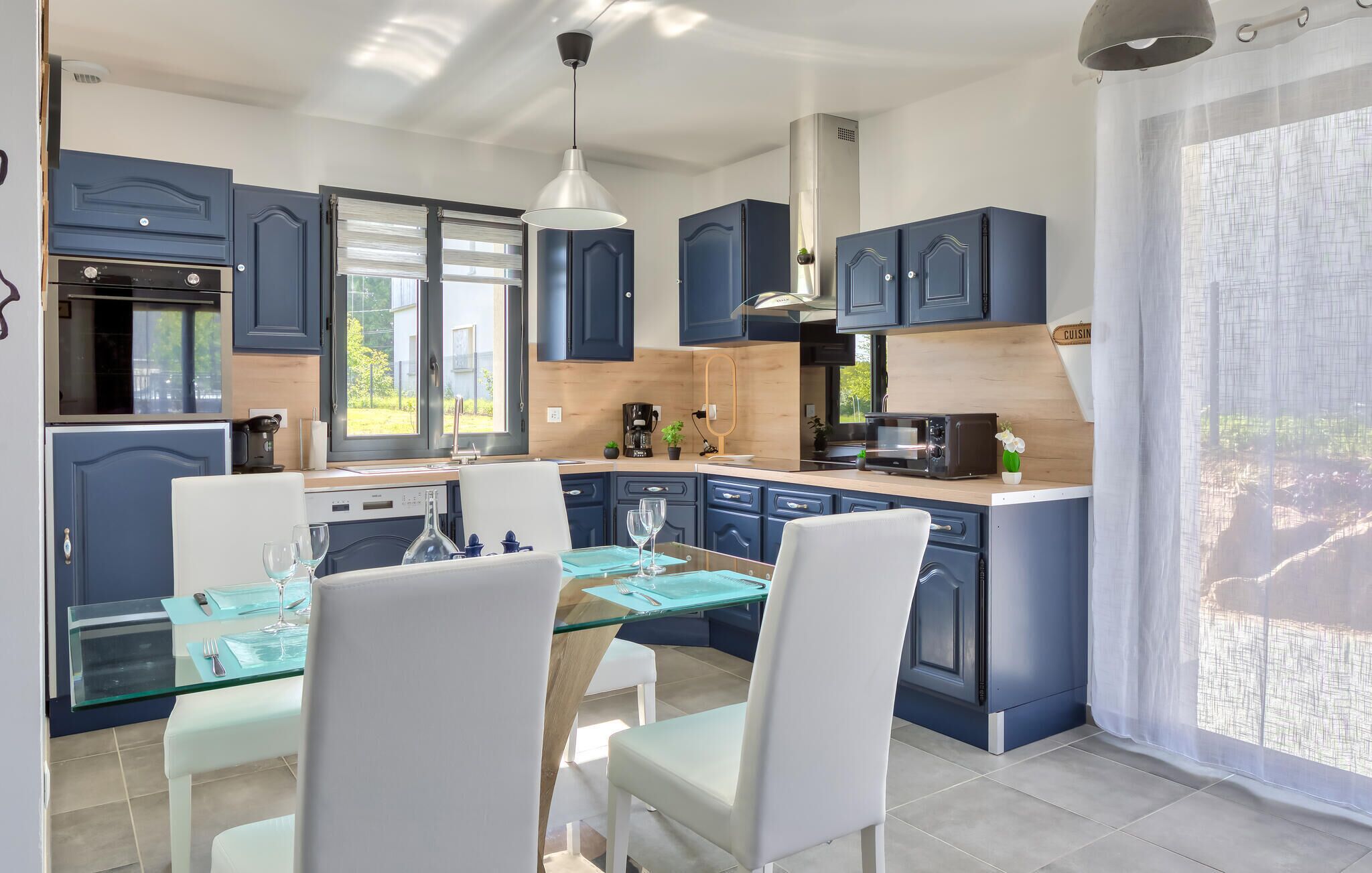 Une cuisine moderne avec des meubles bleus, un coin repas doté d’une table en verre et une vue sur l’extérieur grâce à de grandes fenêtres.