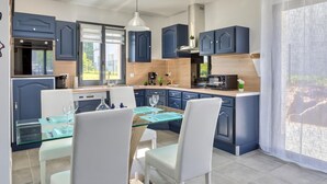 A modern kitchen with blue cabinets, a dining area with a glass table, and a view of the outdoors through large windows.