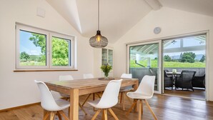 Un comedor con mesa de madera, sillas blancas, una lámpara colgante y ventanas amplias con vistas a la vegetación.
