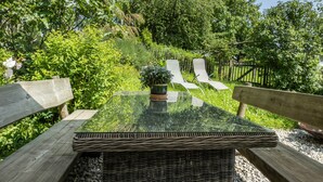 A glass-top table with wicker chairs, surrounded by greenery and a wooden fence.