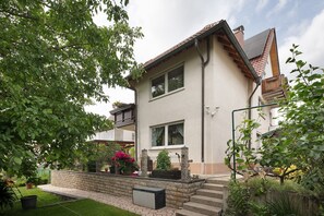 A two-story house with a sloped roof, a brick wall, and a small garden with potted plants and flowers.