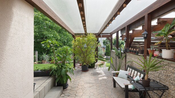 Un patio cubierto con suelo de piedra, un banco de madera, plantas en macetas y una mesa de metal.