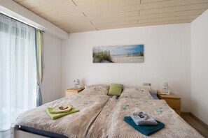 A bedroom with a bed, two bedside tables, a nightstand, a window with curtains, and a beach-themed painting on the wall.