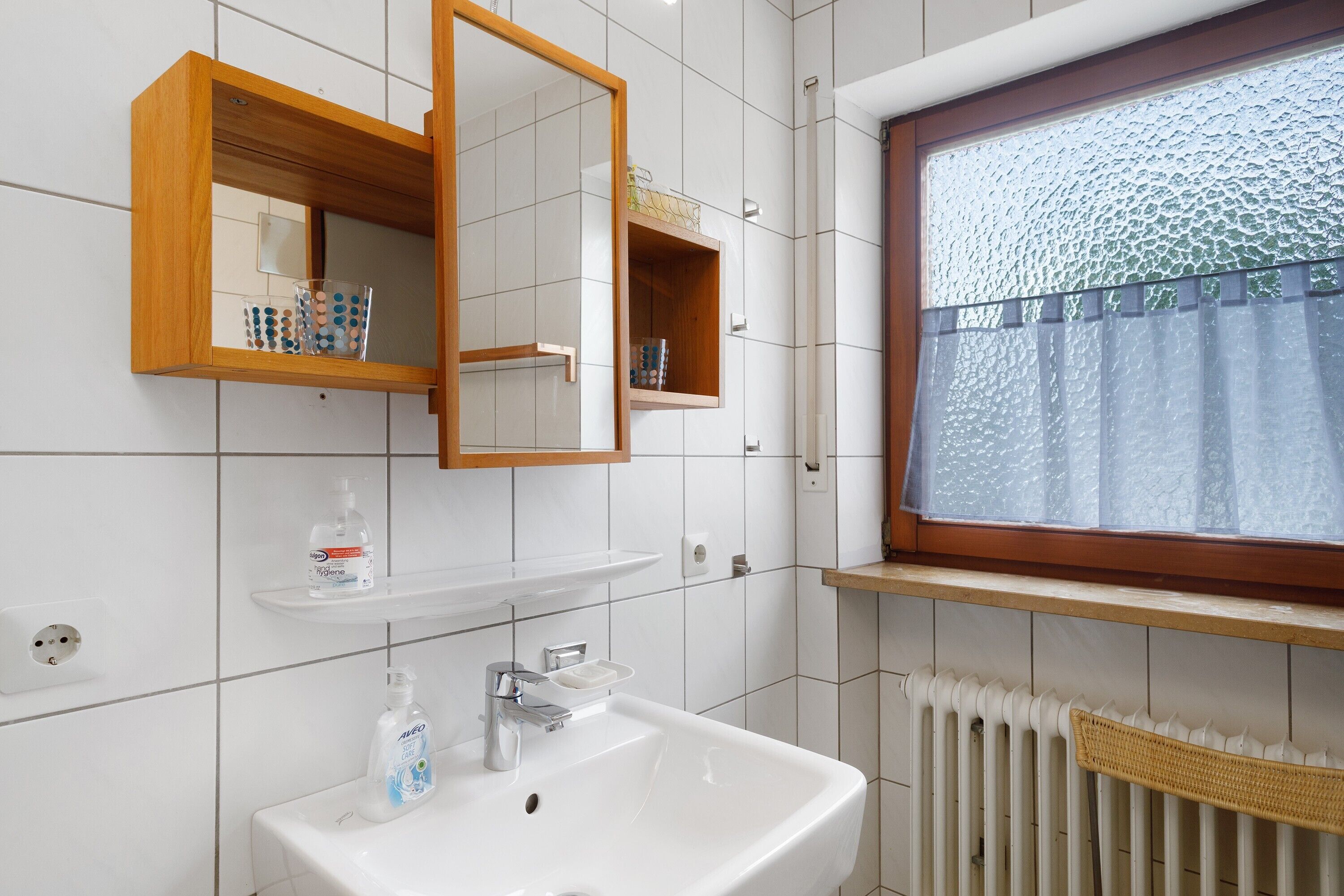 Ein Badezimmer mit weißem Waschbecken, einem Spiegel, einem Fenster mit Milchglas und einer Heizung.