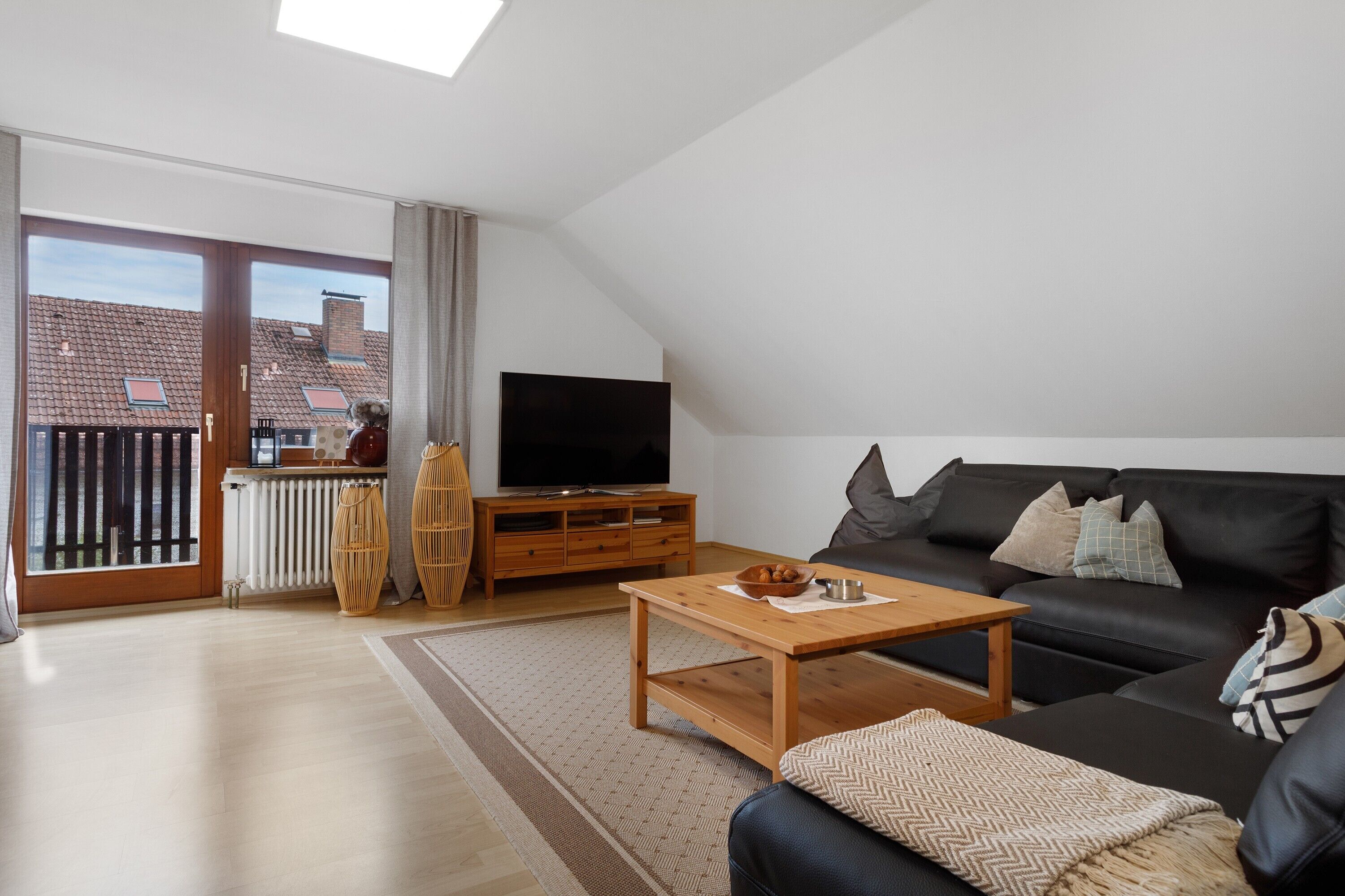 A modern living room with a black leather sofa, a wooden coffee table, a TV on a wooden stand, and a large sliding glass door leading to a balcony.