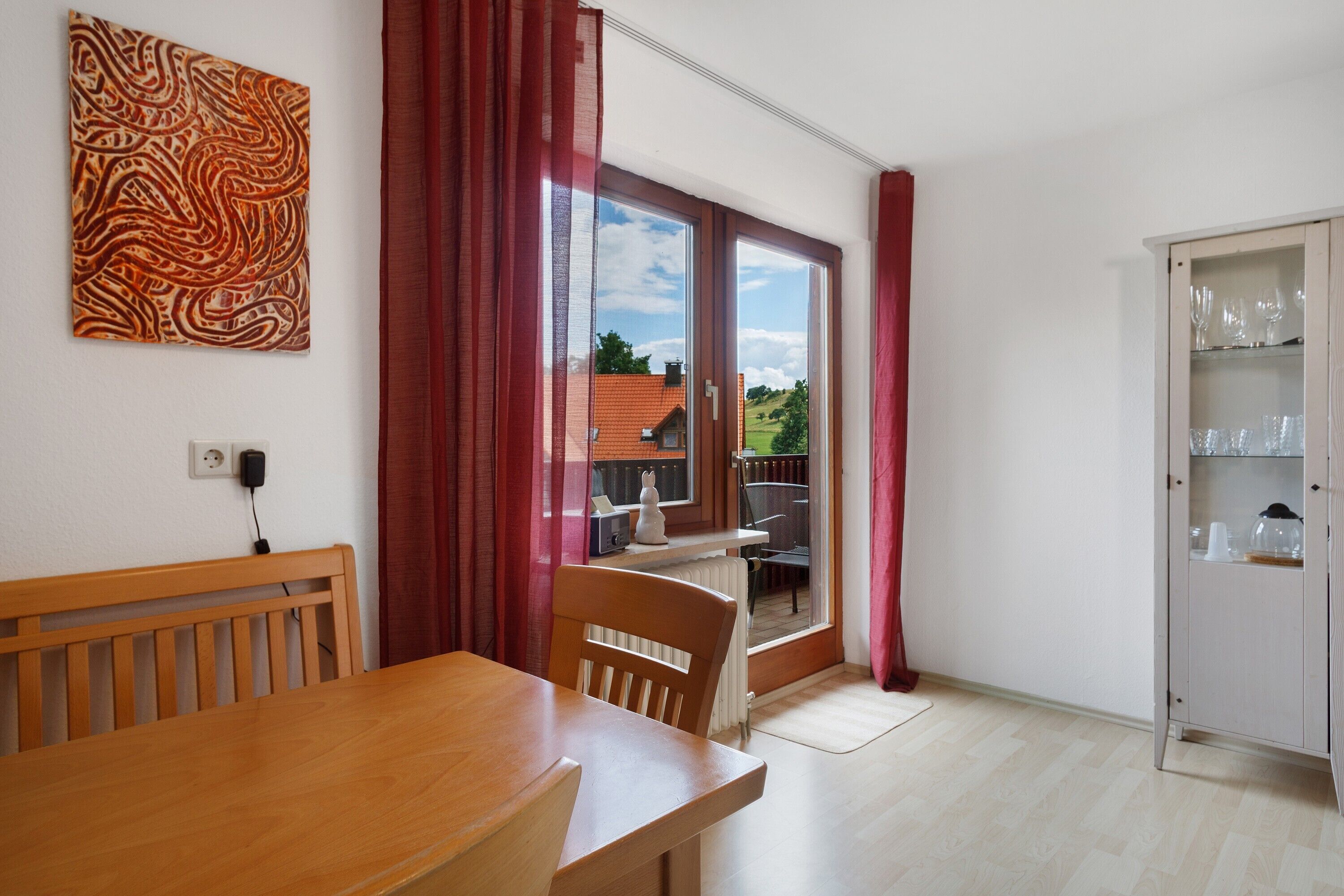 A room with a wooden table, chairs, a radiator, a cabinet with glassware, and a view through a sliding door.