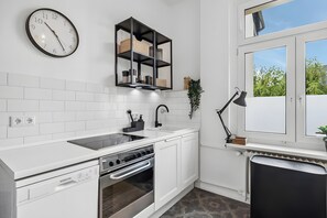 A modern kitchen with white cabinetry, a built-in oven, a dishwasher, a black countertop, a black-framed shelf with baskets, a clock, a window with a view of greenery, and a black trash bin. - Apartment 'Top Wohnung-neu-20 Min Nach Dus' with Wi-Fi (Wuppertal)