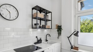Una cocina moderna con armarios blancos, encimera negra, estufa integrada, un lavabo, estante negro con cestas, un reloj y una ventana con vistas a follaje.