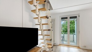 A modern living room with a TV on a wooden stand, a white staircase, and a sliding glass door leading to an outdoor balcony.