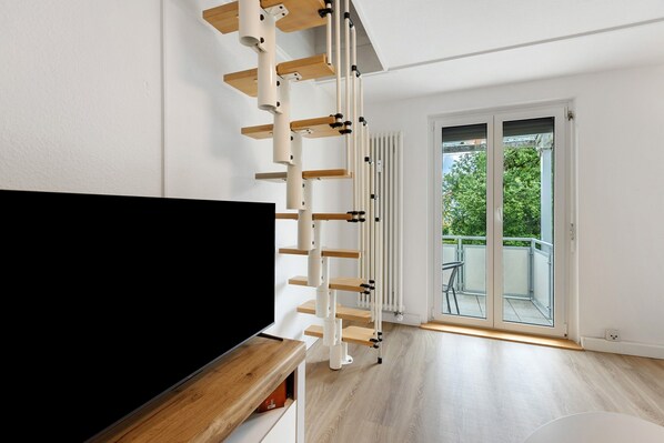 A modern living room with a TV on a wooden stand, a white staircase, and a sliding glass door leading to an outdoor balcony.