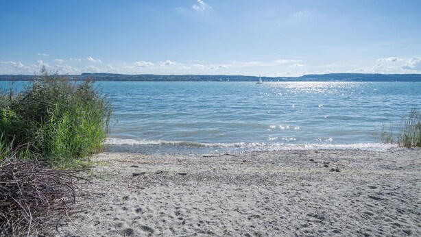 A beach with clear water, a distant sailboat, and a hilly landscape.