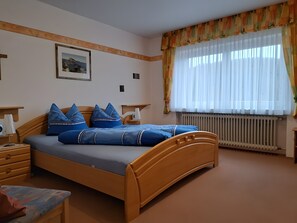 Et soveværelse med en træseng, blåt sengetøj, et natbord med en lampe, et billede i ramme på væggen og et vindue med lette gardiner.