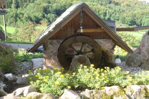 A rustic water mill with a stone base and wooden structure, surrounded by a garden with yellow flowers and a backdrop of a forested hillside.