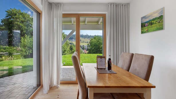 A dining area with a wooden table, chairs, and a view through large windows.