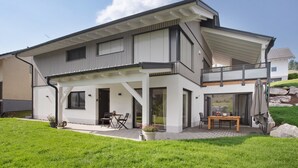 A modern two-story house with a covered patio, a dining area, and a view of the surrounding landscape.