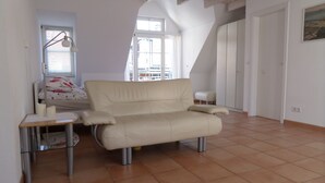 Ein Zimmer mit einem beigen Sofa, einem kleinen Tisch, einer Lampe und einem Fenster mit Jalousien.
