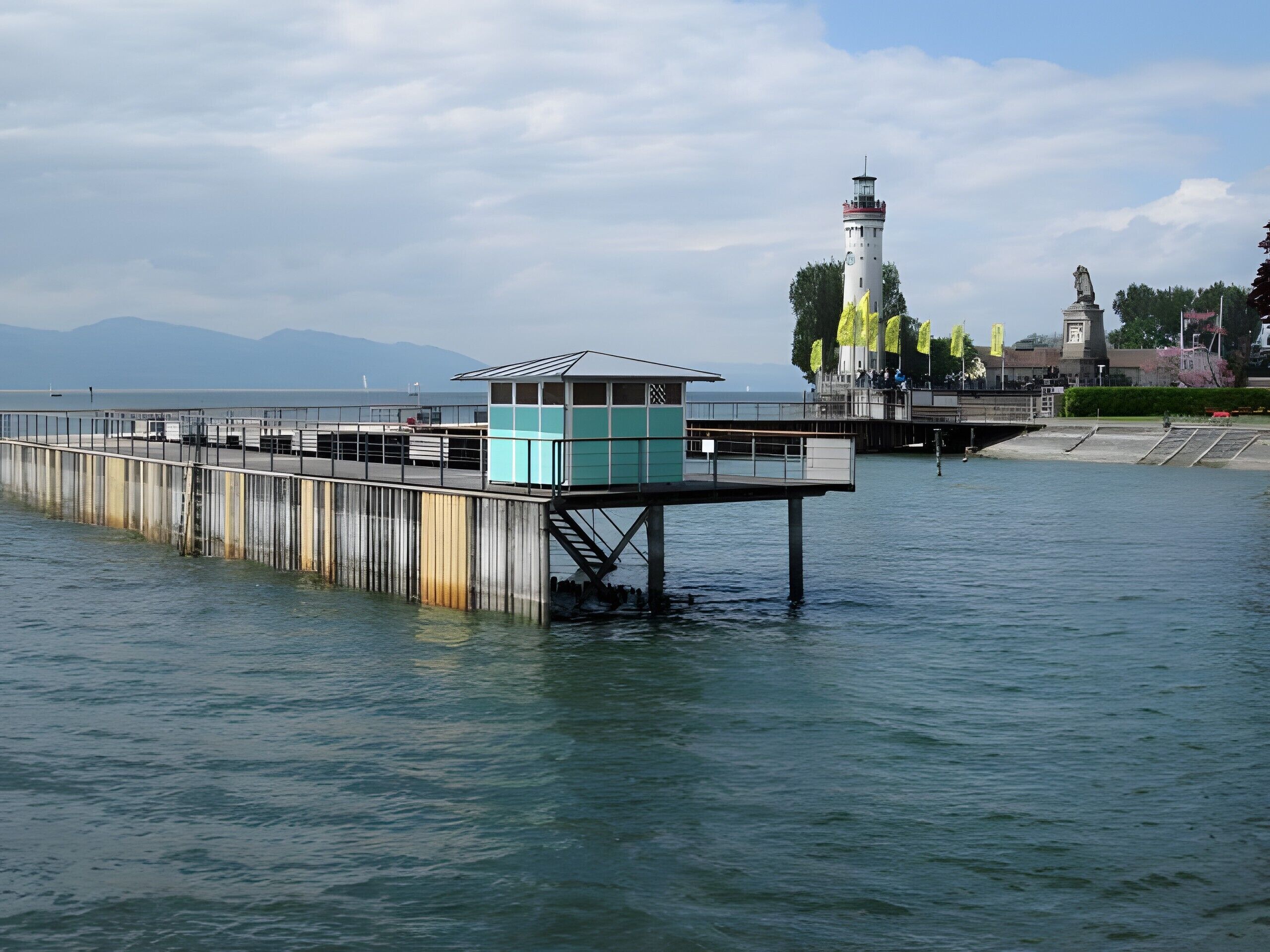 A pier extending into calm water with a lighthouse in the background and a statue on a platform.