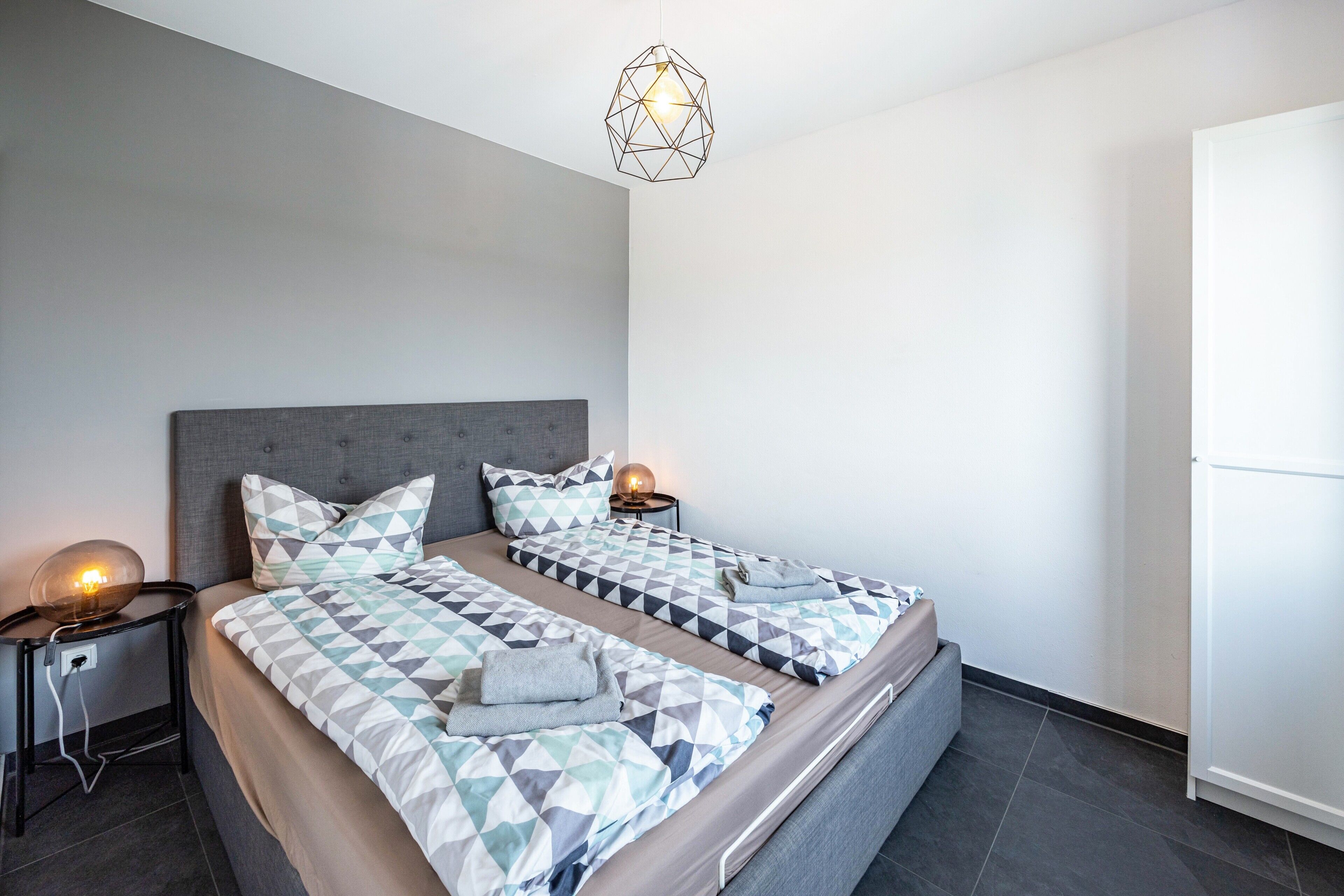 Ett modernt sovrum med två sängar, sängkläder med geometriska mönster, sänglampor och en hänglampa med geometrisk design.
