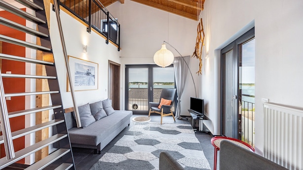 A modern living room with a staircase, a sofa, a chair, a TV, and a large window with a view. - Apartment 'Sonnenwind Am Hainer See' with Lake View and Wi-Fi (Neukieritzsch)