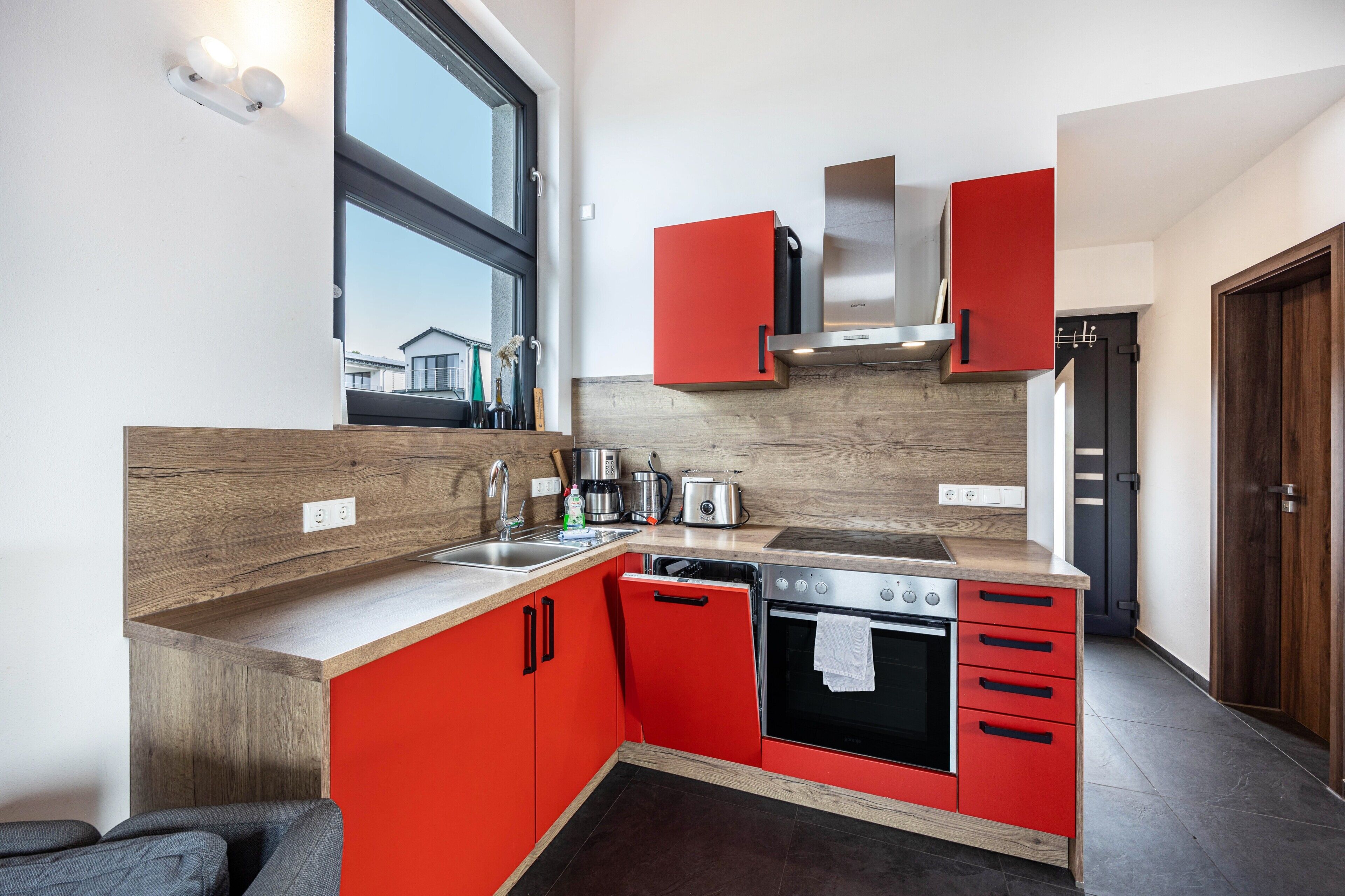 Een moderne keuken met rode kastjes, een roestvrijstalen oven en een raam dat voor natuurlijk licht zorgt.
