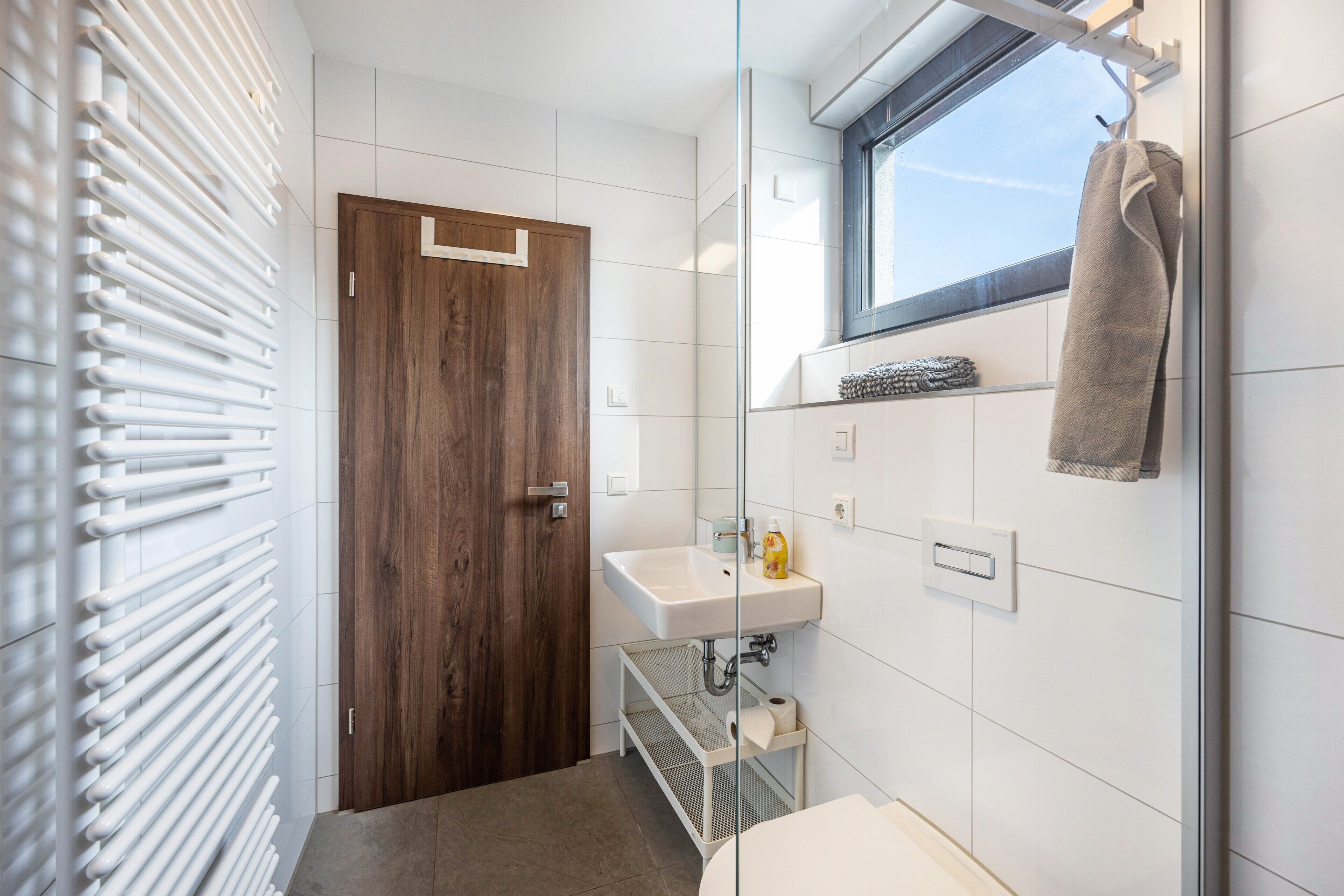 A modern bathroom with a wooden door, a white sink, a towel rack, and a window.