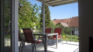 Un espacio al aire libre cubierto con una mesa y sillas, un ventanal de vidrio y vistas a un parque infantil y casas.