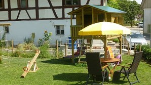 Une structure de jeu en bois, dotée d’un auvent jaune, d’une balançoire et d’une table avec des chaises, installée dans une cour verdoyante.