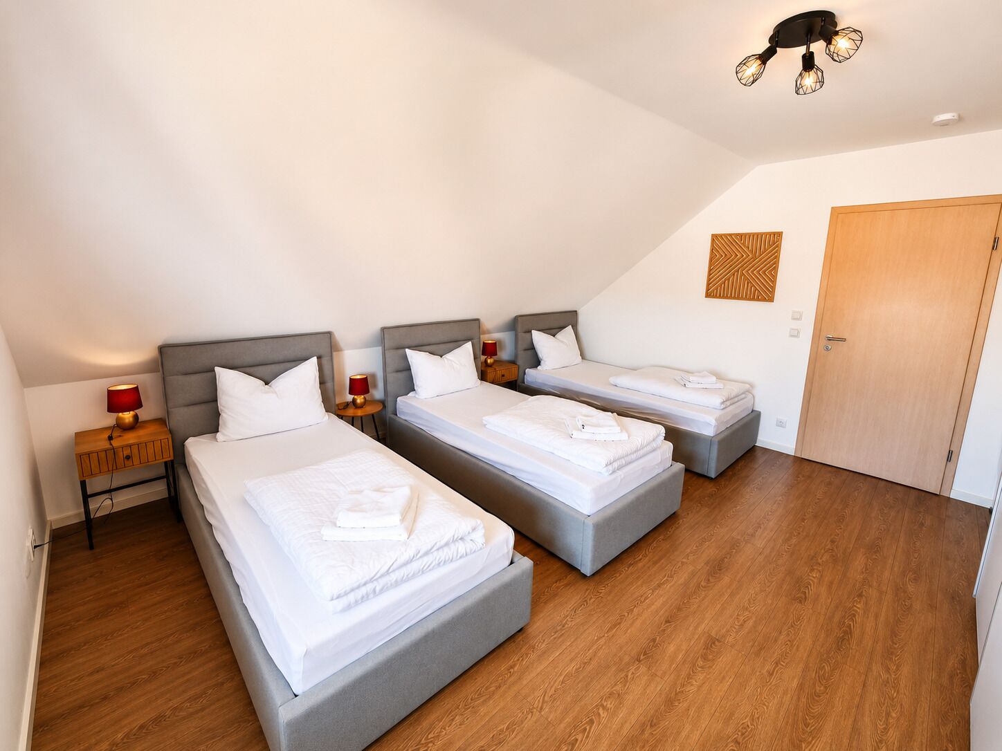 Ein Hotelzimmer mit drei Betten, Holzboden, einer Deckenleuchte und einer Holztür.