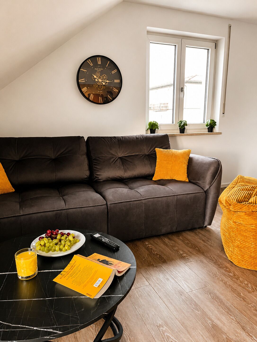 Un salon avec un canapé marron, une table basse ronde, une horloge et une fenêtre ornée de plantes.