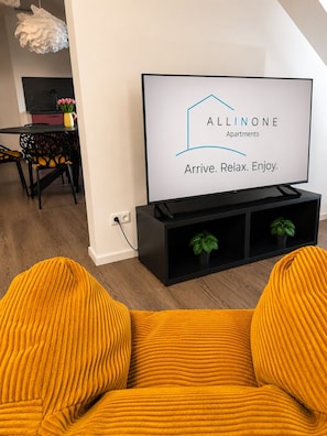 Een moderne woonkamer met een flatscreen televisie waarop 'Allinone Apartments' staat en de slogan 'Arrive. Relax. Enjoy.'