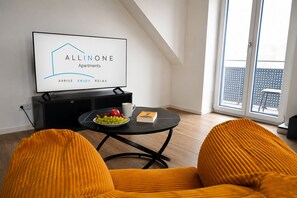A modern living room with a black TV displaying a logo, a round table with fruit and a book, and yellow upholstered chairs.