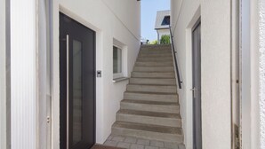 Una escalera estrecha que sube a una puerta con un ventanal arriba, un felpudo en la parte inferior y una ventana pequeña en la pared.