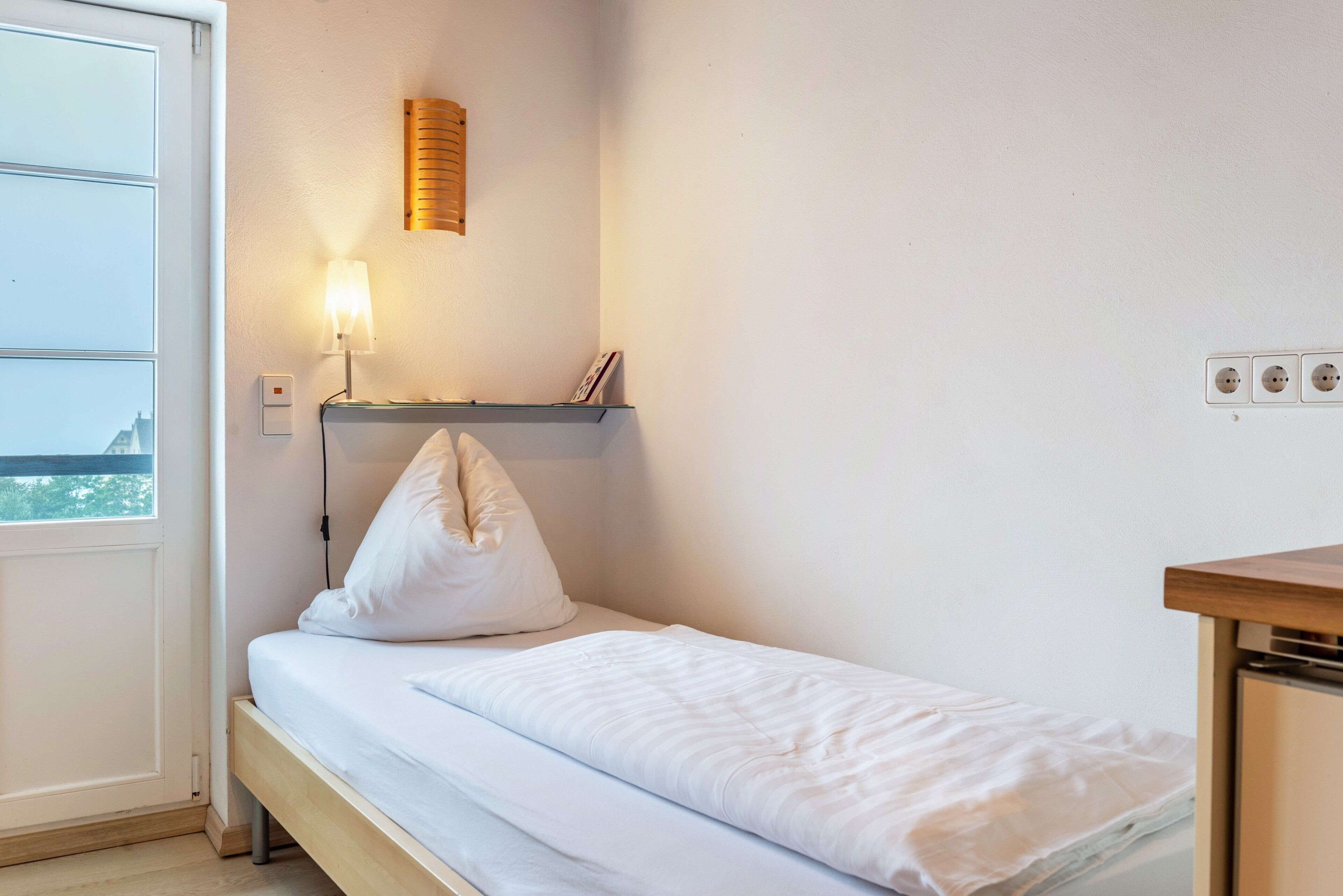 Un lit simple avec une literie blanche, une lampe de chevet, une étagère avec un cadre photo et une fenêtre donnant sur un paysage.