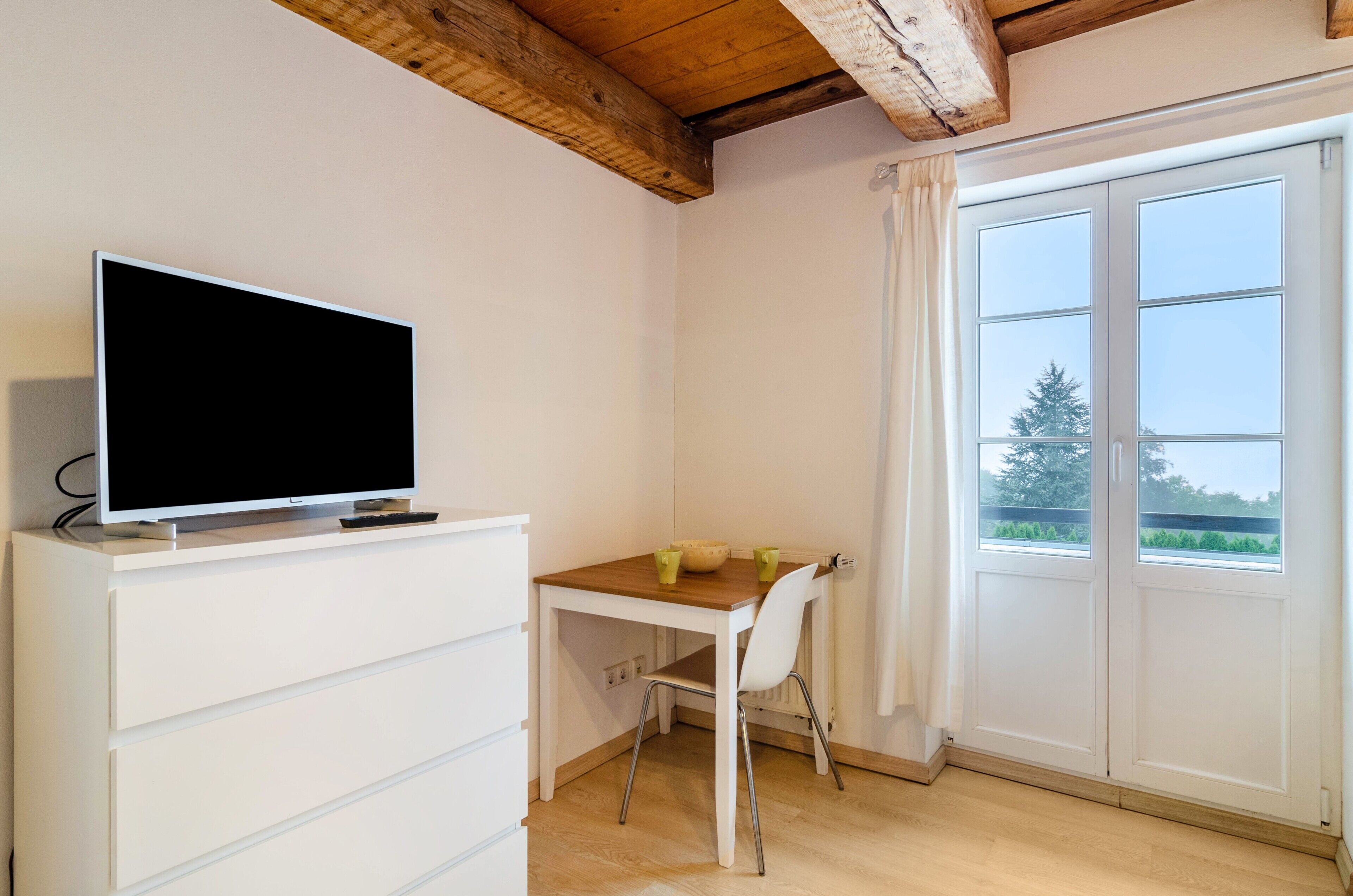 A room with a TV on a white dresser, a wooden table with two chairs, and a large window with curtains.