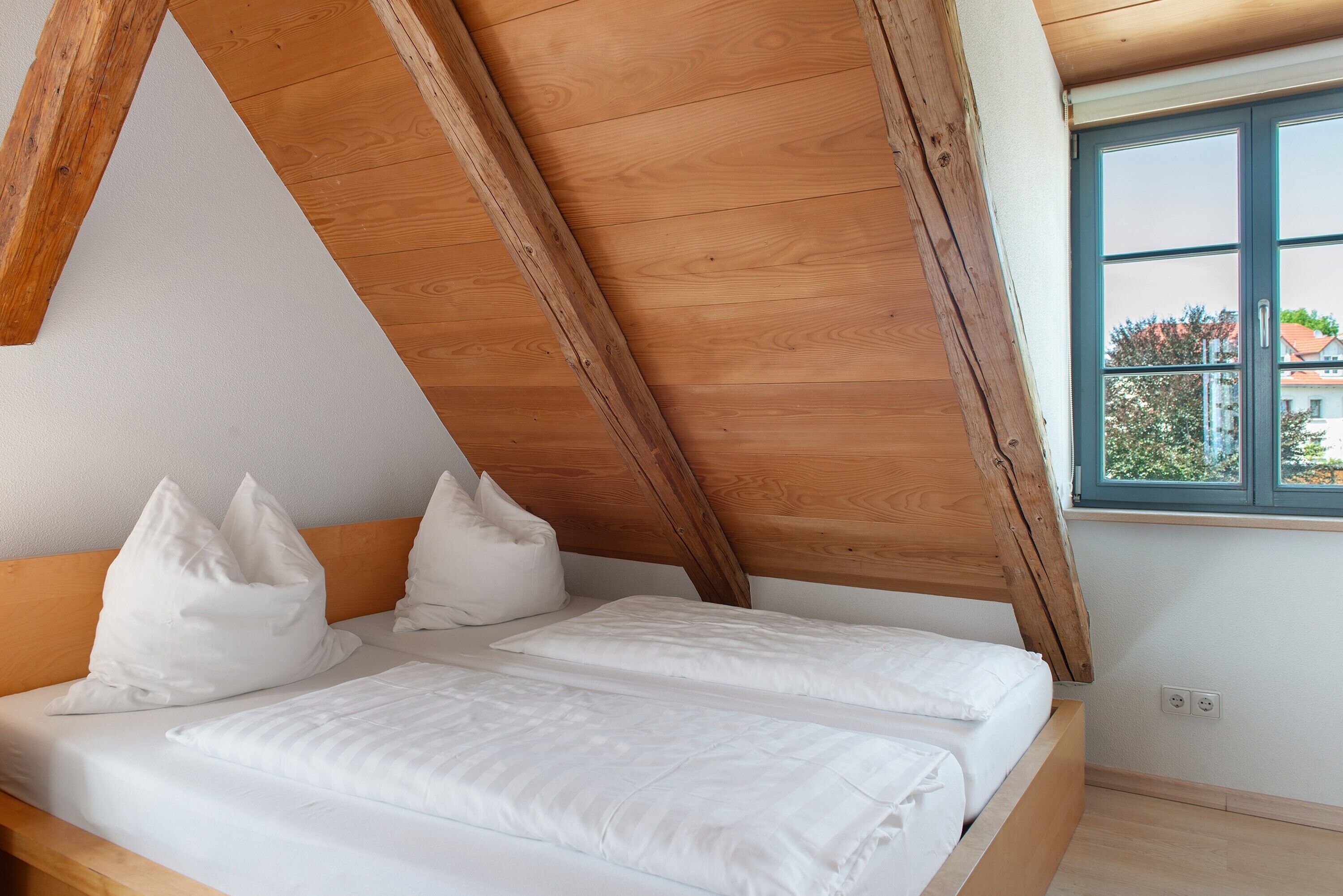 A bedroom with a sloped ceiling, wooden headboard, white bedding, and a window with a view of buildings.