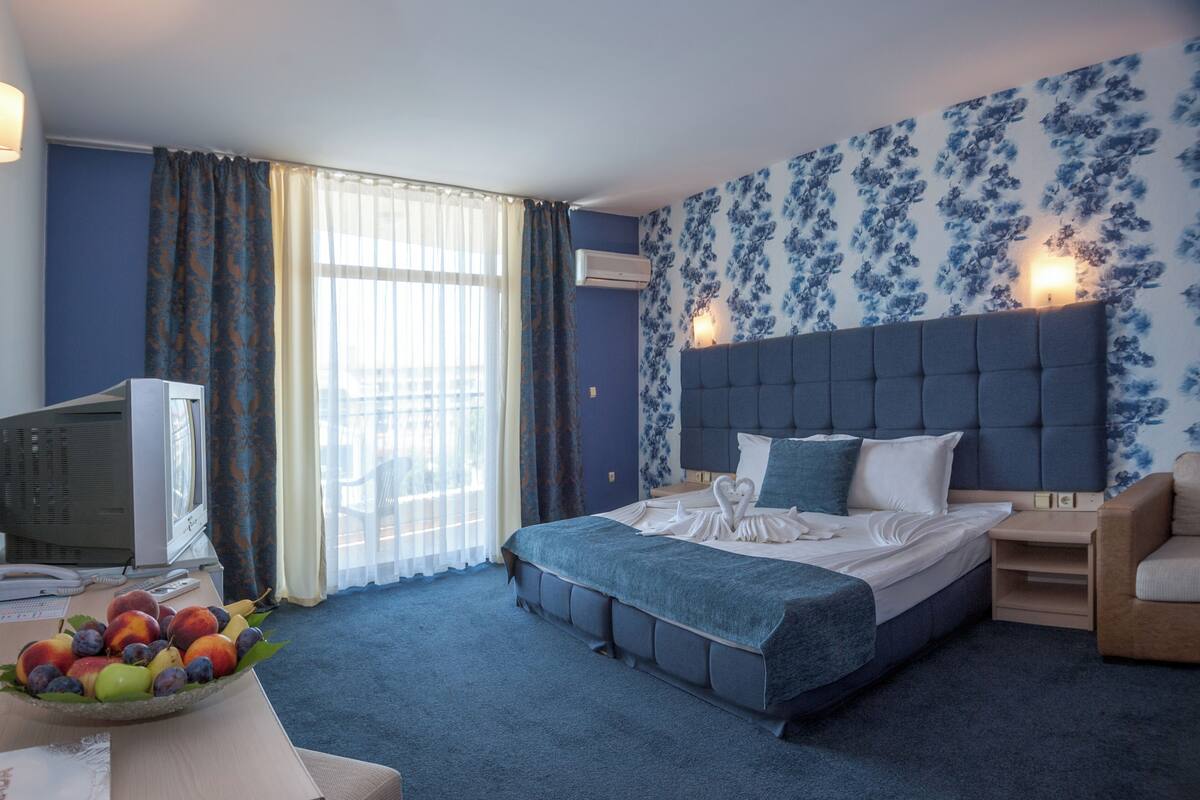 A hotel room with a large bed, a desk with a computer, a fruit bowl, and a view of the city through the window.
