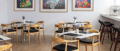 A dining area with wooden chairs and tables, framed artworks on the wall, and a white counter with a vase.